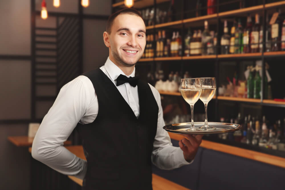 young waiter smiling while holding tray with wine glasses in the restaurant