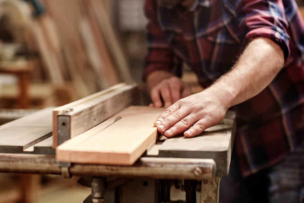 cropped hands of craftsman cutting wooden plank with circular saw in workshop