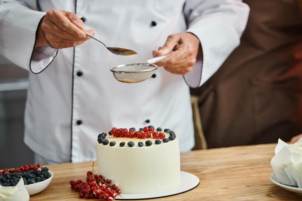 A male pastry chef using a sifter to add decorations to a newly baked cake